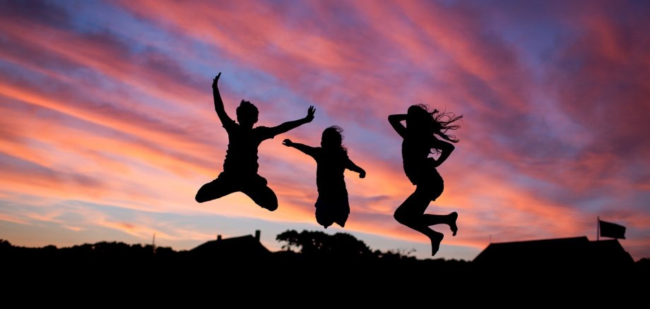 Drei Personen springen in die Höhe. Man erkennt nur die Silhouetten, im Hintergrund Sonnenuntergang